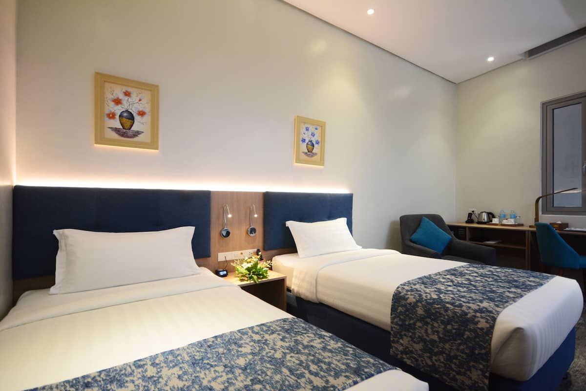 A hotel room with two beds, a desk, a chair, and two framed artworks on the wall.