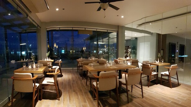 A modern dining area with wooden tables and chairs, large windows offering a city view, and a ceiling fan. - Blue Lotus Hotel Davao (Davao)