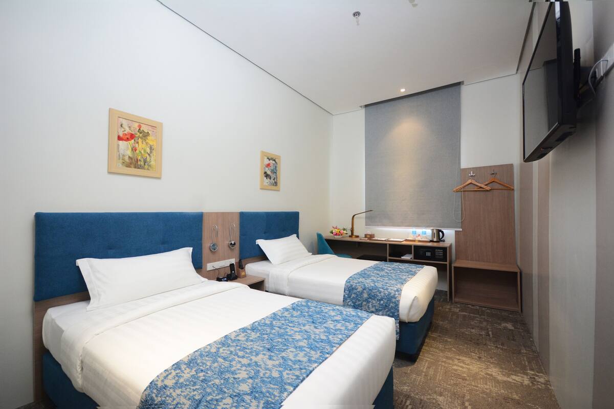 A hotel room with two beds, a desk with a chair, a TV mounted on the wall, and artwork on the walls.