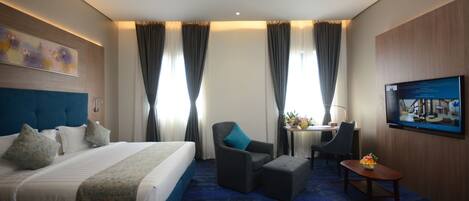 A hotel room with a large bed, a desk with a chair, a small table with a vase, and a television on the wall.