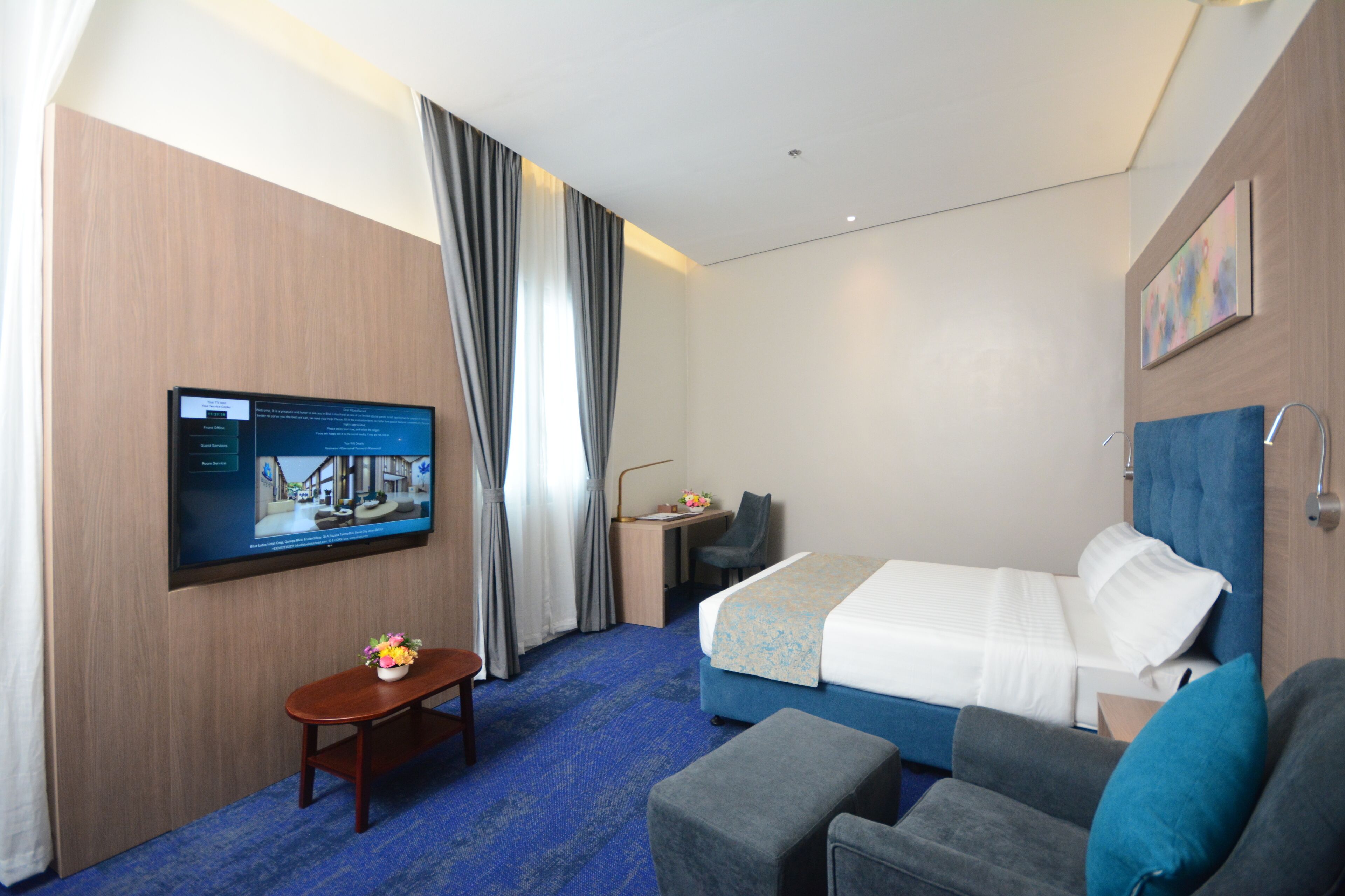 A hotel room with a bed, a desk, a TV, and a small table with flowers.