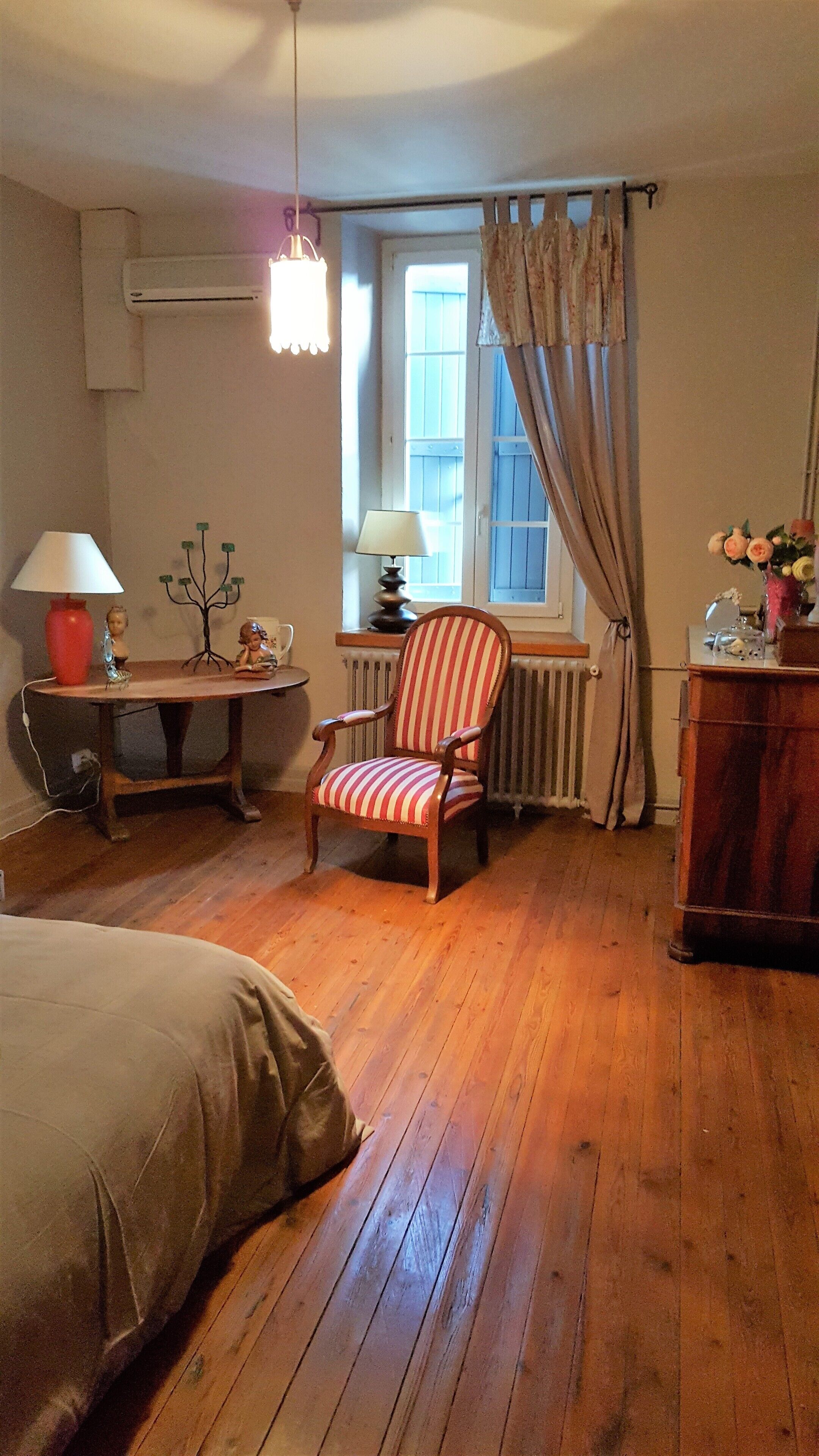 Une pièce avec un plancher en bois, un fauteuil à rayures, une table avec une lampe, un radiateur et une fenêtre avec des rideaux.