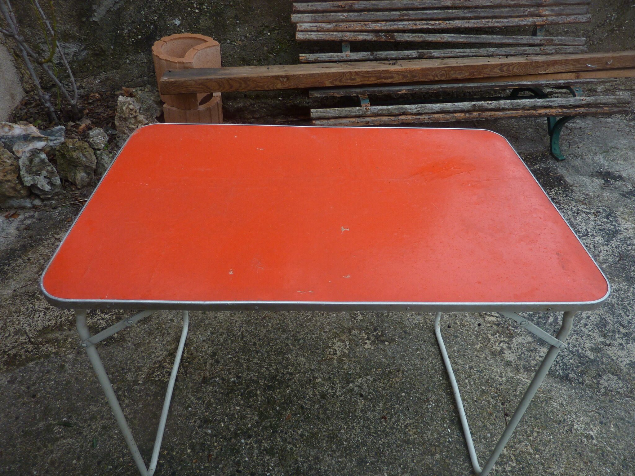 A red metal table with a metal frame, placed outdoors on a gravel surface.