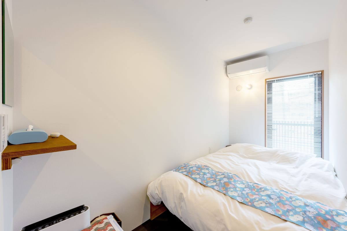 Un dormitorio con una cama, un estante con un dispensador de pañuelos, un radiador y una ventana con persianas.