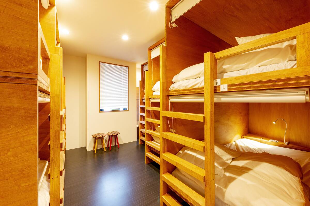 Habitación con literas de madera, una ventana con persianas, un taburete pequeño y un escritorio con lámpara.