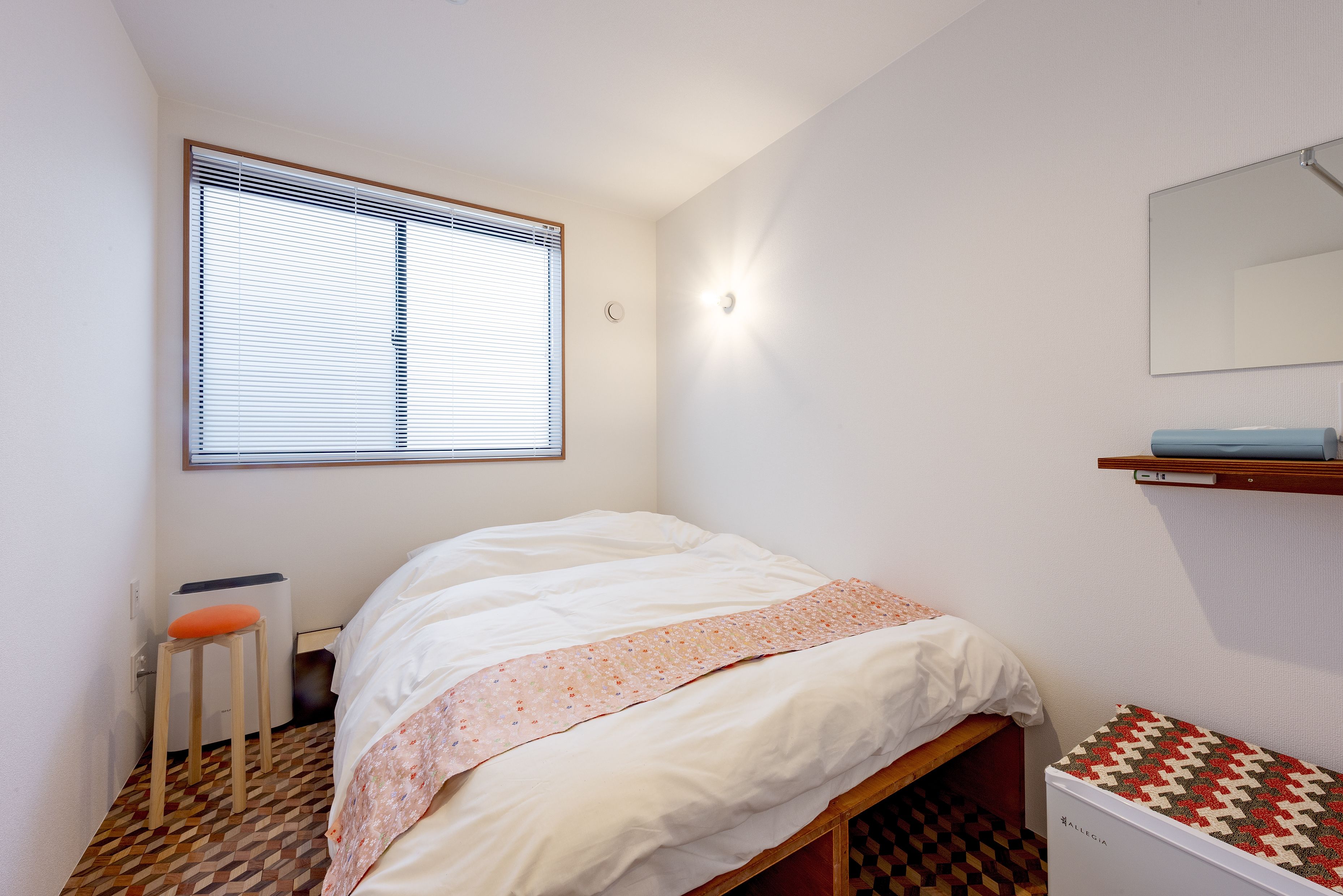 A small, clean room with a bed, a stool, a radiator, and a mirror on the wall.