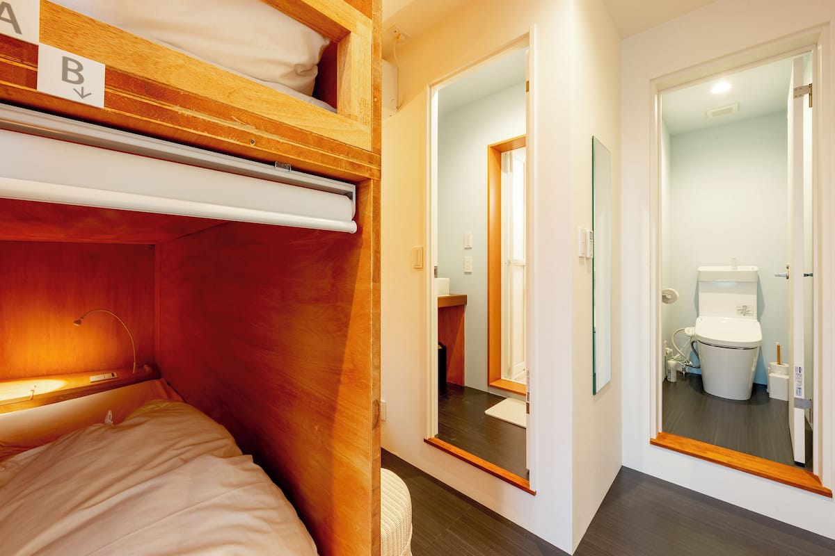 Una cama litera con cabecera de madera, un espejo y una puerta que da a un baño con inodoro y lavamanos.