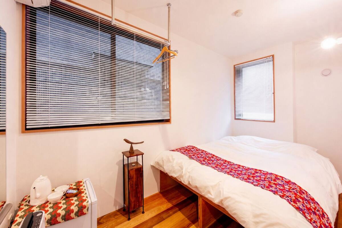 Un dormitorio con cama, suelo de madera, ventana con persianas, mesita de noche con hervidor y un estante con objetos decorativos.