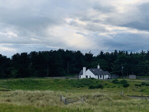 Ein weißes Haus mit Giebeldach, umgeben von einem eingezäunten Feld und dichtem Wald.