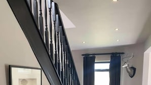 Una escalera con barandillas negras, un cuadro enmarcado en la pared, un florero con flores sobre una consola y un sofá de cuero marrón con cojines.