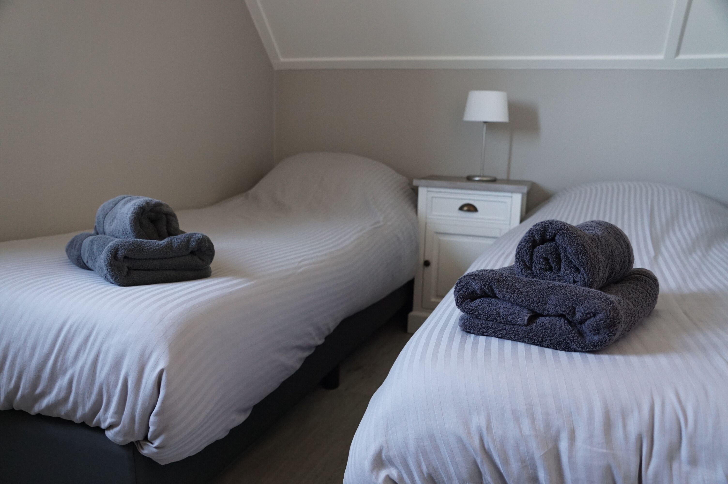 Une chambre d’hôtel avec deux lits, chacun agrémenté d’une serviette pliée. On y trouve une table de chevet avec une lampe et un tiroir.