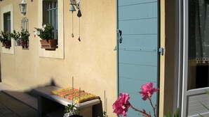 Ein Gebäude mit beige-farbener Fassade, einer blauen Tür und einer Bank mit einem farbenfrohen Kissen. Es gibt Blumentöpfe und eine hängende Leuchte.