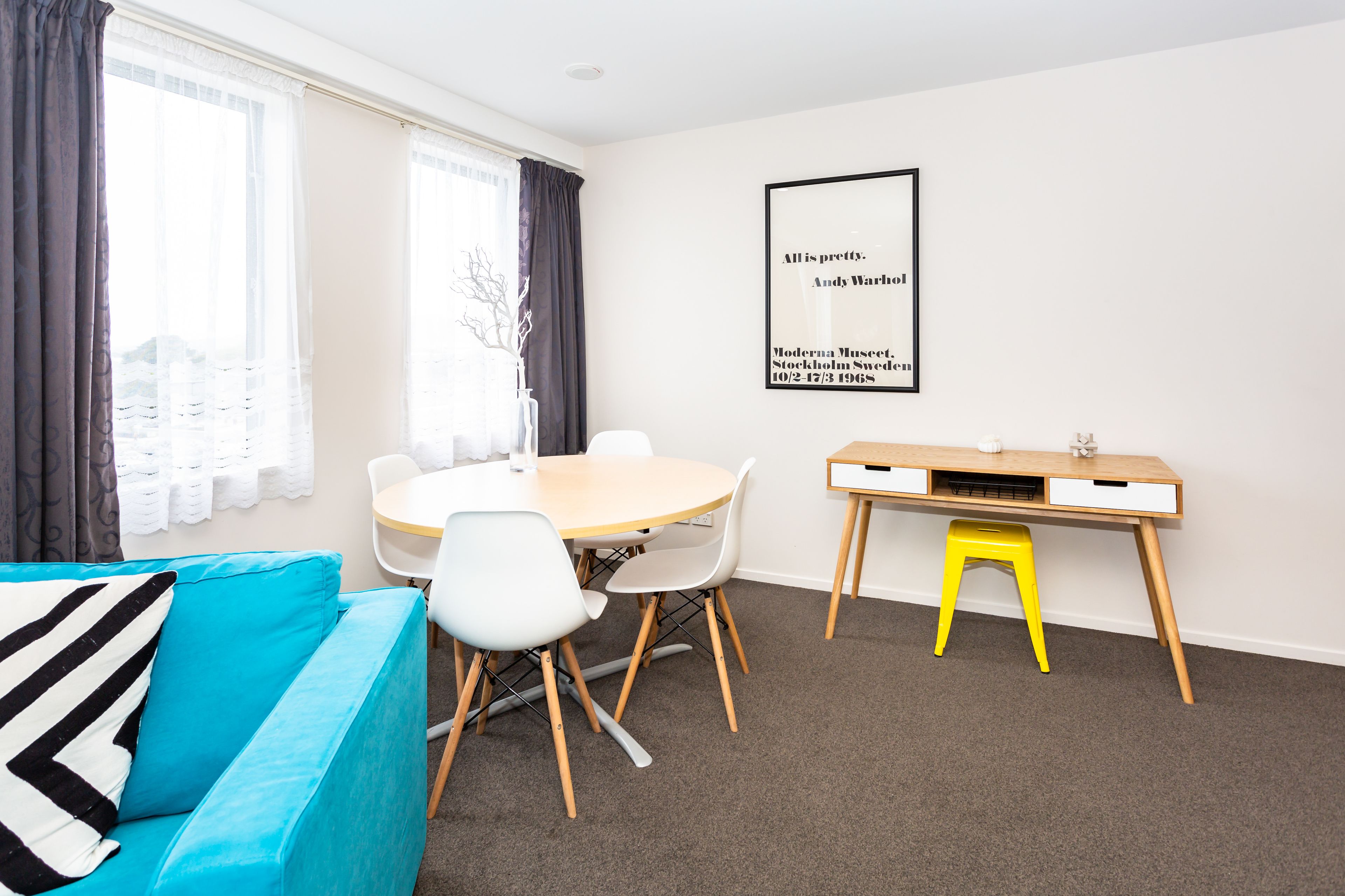 Une pièce moderne avec une table ronde, des chaises, un canapé bleu, un tabouret jaune et un tableau encadré au mur.