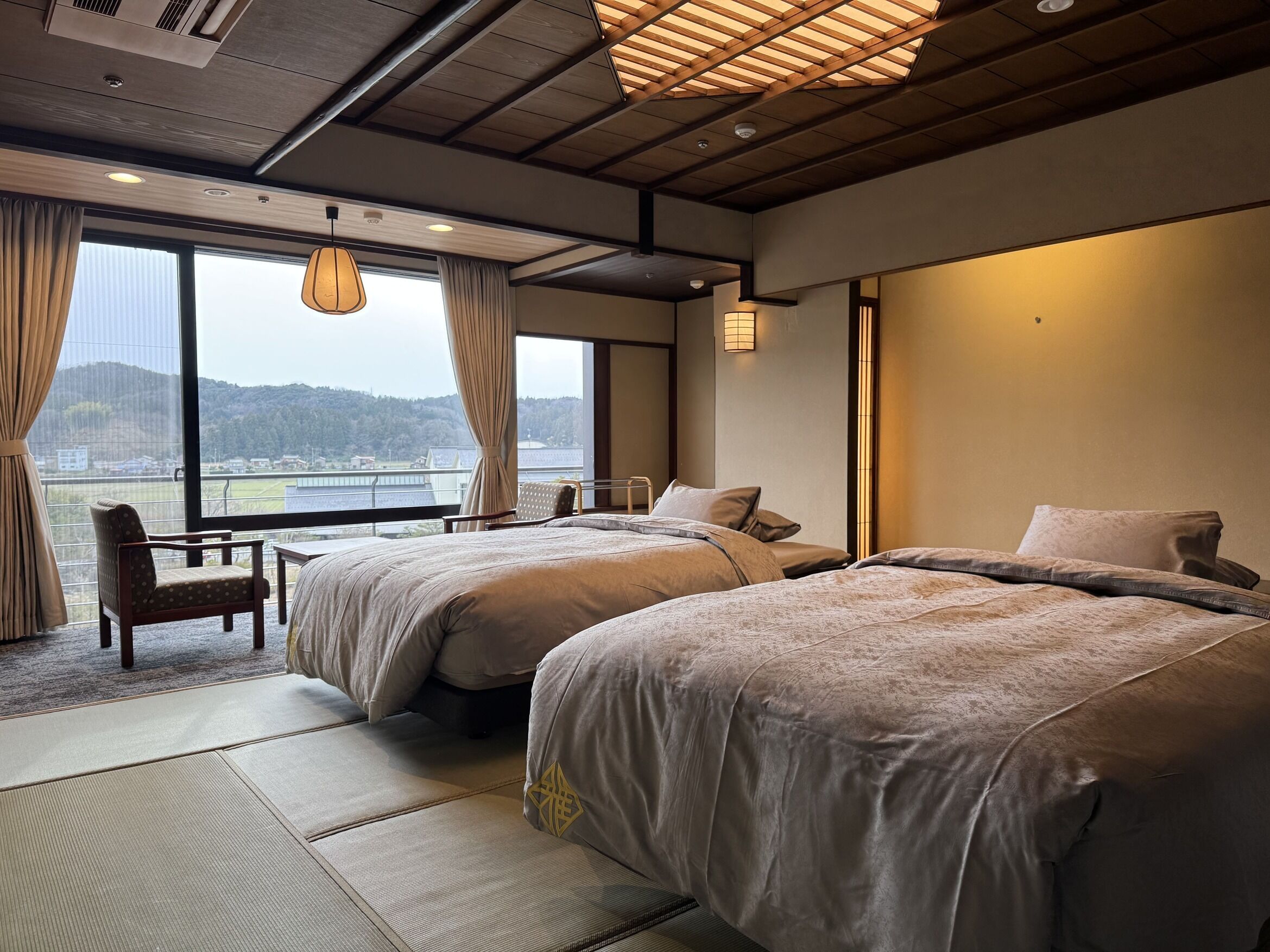 Quarto de hotel com duas camas, uma cadeira, uma mesinha e vista para a água.