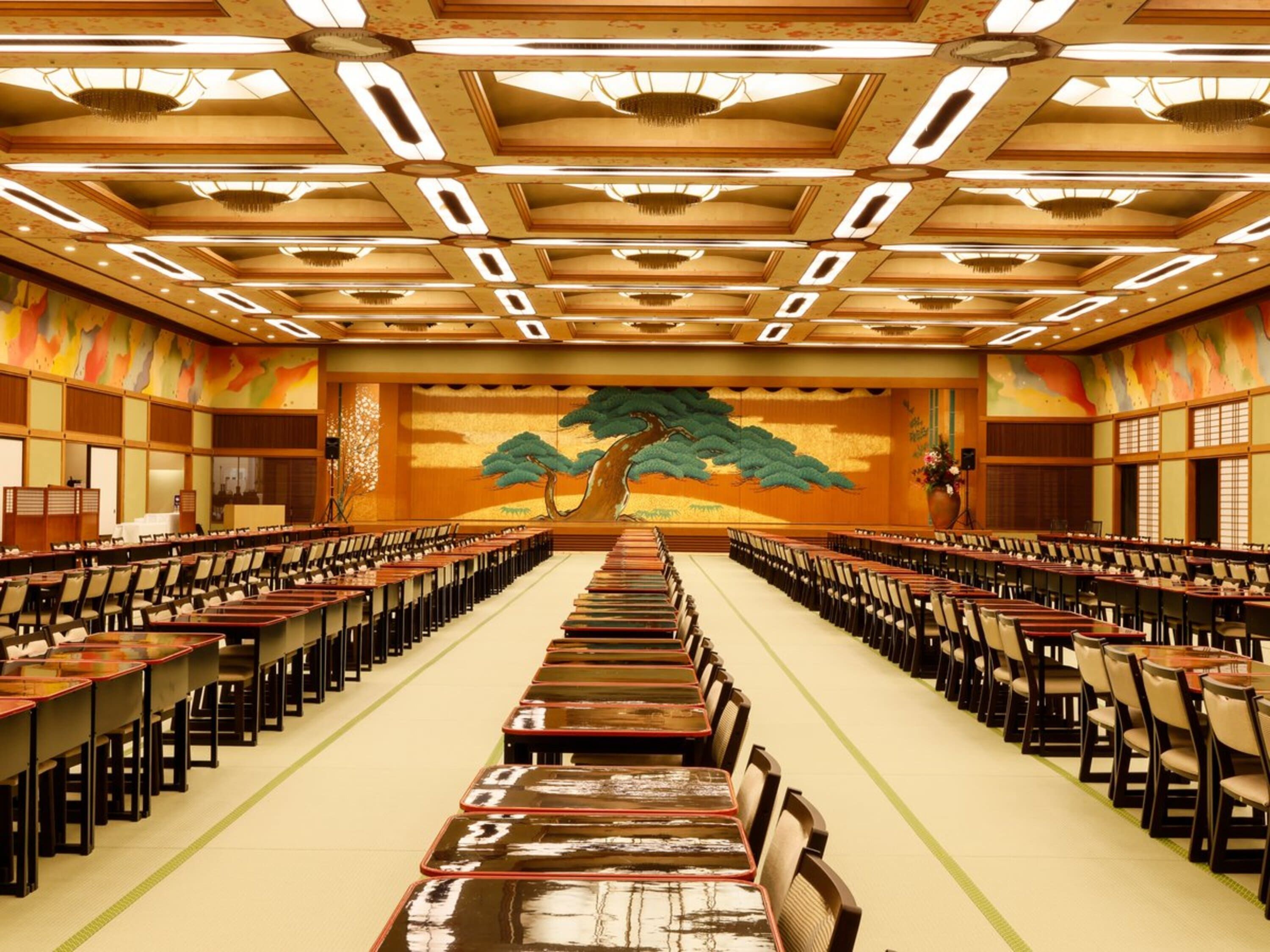 Une salle de conférence avec des rangées de tables et de chaises, une grande fresque sur le mur du fond et un plafond doté d’un éclairage encastré.