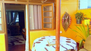 A room with a round table, a chair, a bed, a window with a grid pattern, and a wall-mounted decorative item.