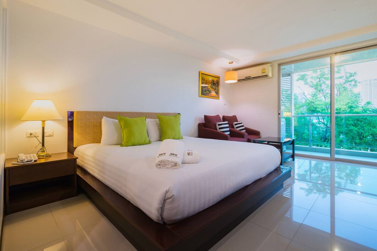 Une chambre d’hôtel avec un grand lit, une table de chevet avec une lampe, une chaise et une vue sur la verdure depuis un balcon.