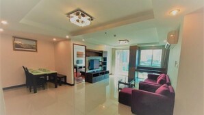 A modern living room with a flat-screen TV, a dining table with chairs, a sofa, and a large window with a view. - Nice Residence Hotel Bangkok (Bangkok)