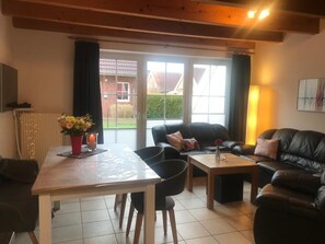 A living room with a dining table, two black chairs, a black leather sofa, and a wooden coffee table. There is a window with curtains, a radiator, and a vase with flowers on the table. -  renovated in 2024 !!! very cozy and comfortable, 3 bedrooms, WLAN, all incl. (Wangerland)