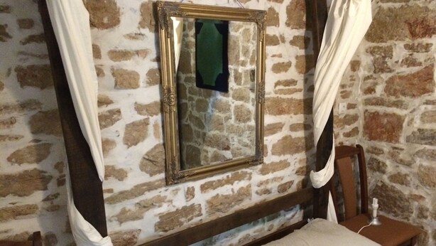 A room with a stone wall, a bed with white bedding, a wooden headboard, a nightstand with a lamp, and a framed mirror.