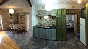 A compact kitchen with a dining area, stone walls, and a green door leading to a bedroom.