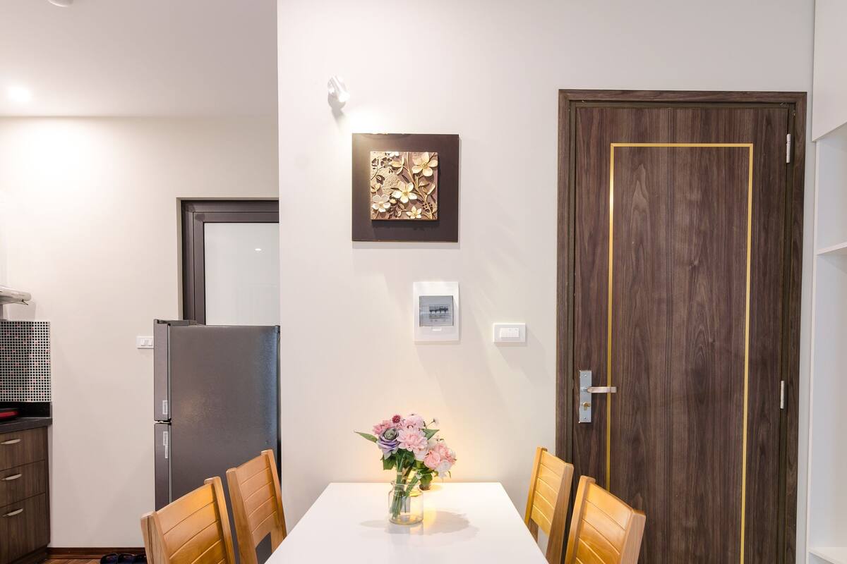 Un coin repas avec une table blanche et des chaises en bois, un vase rempli de fleurs et, au fond, une porte en bois.