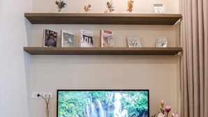 Un téléviseur à écran plat est posé sur un meuble télé en bois doté de trois tiroirs. Au-dessus, on trouve deux étagères ornées d’objets décoratifs et de photos encadrées.