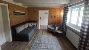 A cozy room with a dark grey sofa, a grey armchair, and a grey rug. There is a wooden wall with a door and a window with checkered curtains.