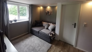 A cozy room with a grey sofa, a window with checkered curtains, a small table with a lamp, and a door.