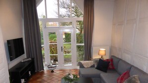 Ein Wohnzimmer mit grauem Sofa, einem schwarzen Couchtisch, einem Fernseher auf einem schwarzen Schrank und einem großen Fenster mit Vorhängen.