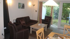 A living room with a brown leather sofa, a wooden coffee table, and a dining table with chairs. There is a lamp, a painting on the wall, and a view of the garden through the glass door. - Modern semi-detached house on the North Sea for up to 4 people (Butjadingen)