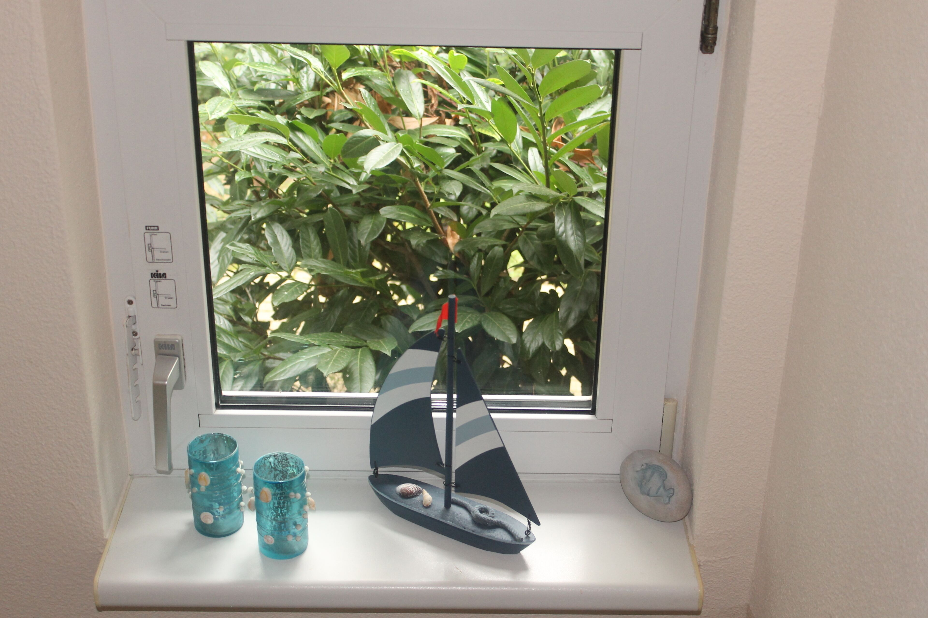 A window with a sailboat model, two decorative cups, and a small stone on a windowsill.