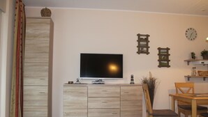 A modern living room with a flat-screen TV, wooden cabinet, dining table with chairs, and decorative wall clocks. - Modern semi-detached house on the North Sea for up to 4 people (Butjadingen)