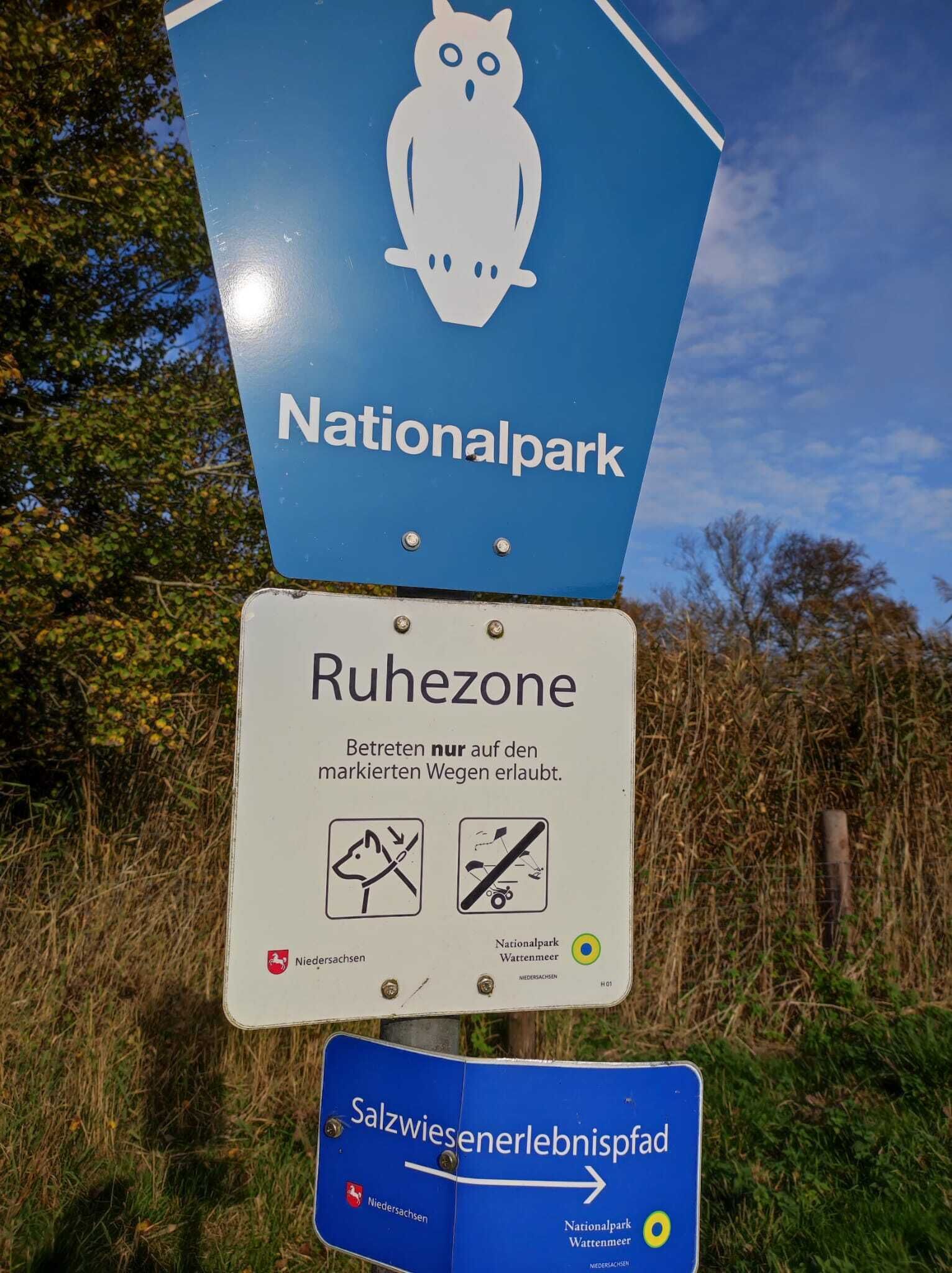 A signpost with multiple signs, including a blue sign with an owl symbol and the text 'Nationalpark' and a white sign with 'Ruhezone' and symbols indicating no dogs or bicycles.