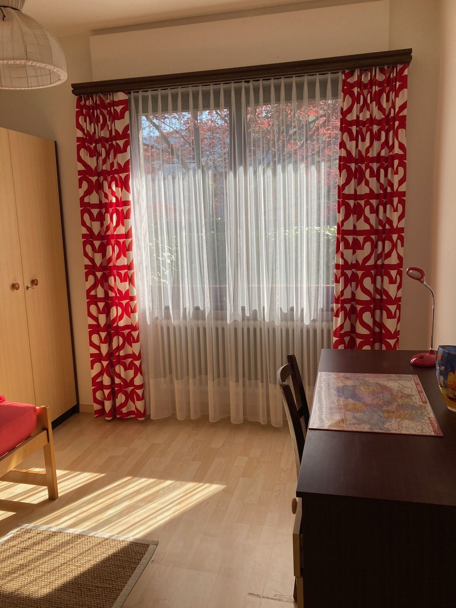 Une pièce avec un plancher en bois, une table à manger ornée d’une carte, une chaise, une armoire en bois et des rideaux légers à motifs rouges.