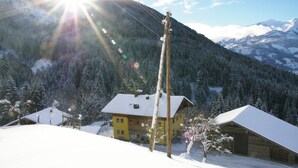 Un edificio amarillo con techo nevado, rodeado de un bosque y montañas al fondo.