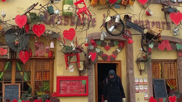 A building with a yellow facade, red shutters, and a clock. There are heart-shaped decorations and various items like a bicycle and a rooster statue. - Magnificent spacious apartment with WIFI, ideal for PMR. 3-star rating. (Xonrupt-Longemer)