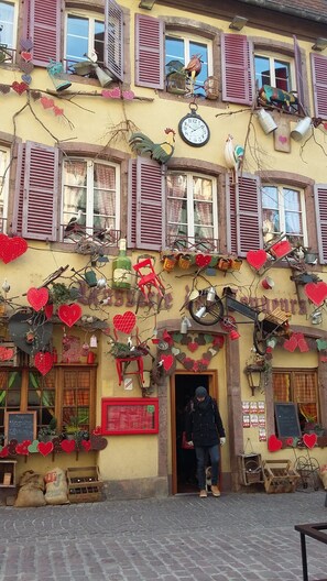 A building with a yellow facade, red shutters, and a clock. There are heart-shaped decorations and various items like a bicycle and a rooster statue. - Magnificent spacious apartment with WIFI, ideal for PMR. 3-star rating. (Xonrupt-Longemer)