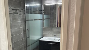 A modern bathroom with a glass shower enclosure, a sink with a mirror, and dark cabinetry. - Rental three-room apartment in Saint Lary (Vignec)