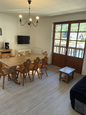 A dining area with a wooden table and chairs, a television mounted on the wall, and a window with a view of the outdoors.