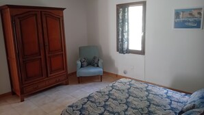 Ein Schlafzimmer mit einem Bett, einem Kleiderschrank, einem Sessel, einem Fenster mit Vorhängen und einem Bild an der Wand.
