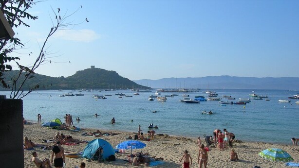 Ein Strand mit badenden Personen, Booten im Wasser und Menschen, die sich sonnen.