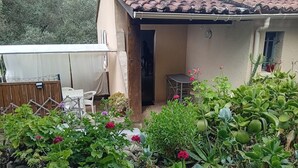 Ein kleines Haus mit Ziegeldach, einer Terrasse mit Tisch und Stühlen sowie ein Garten mit verschiedenen Pflanzen und Blumen.