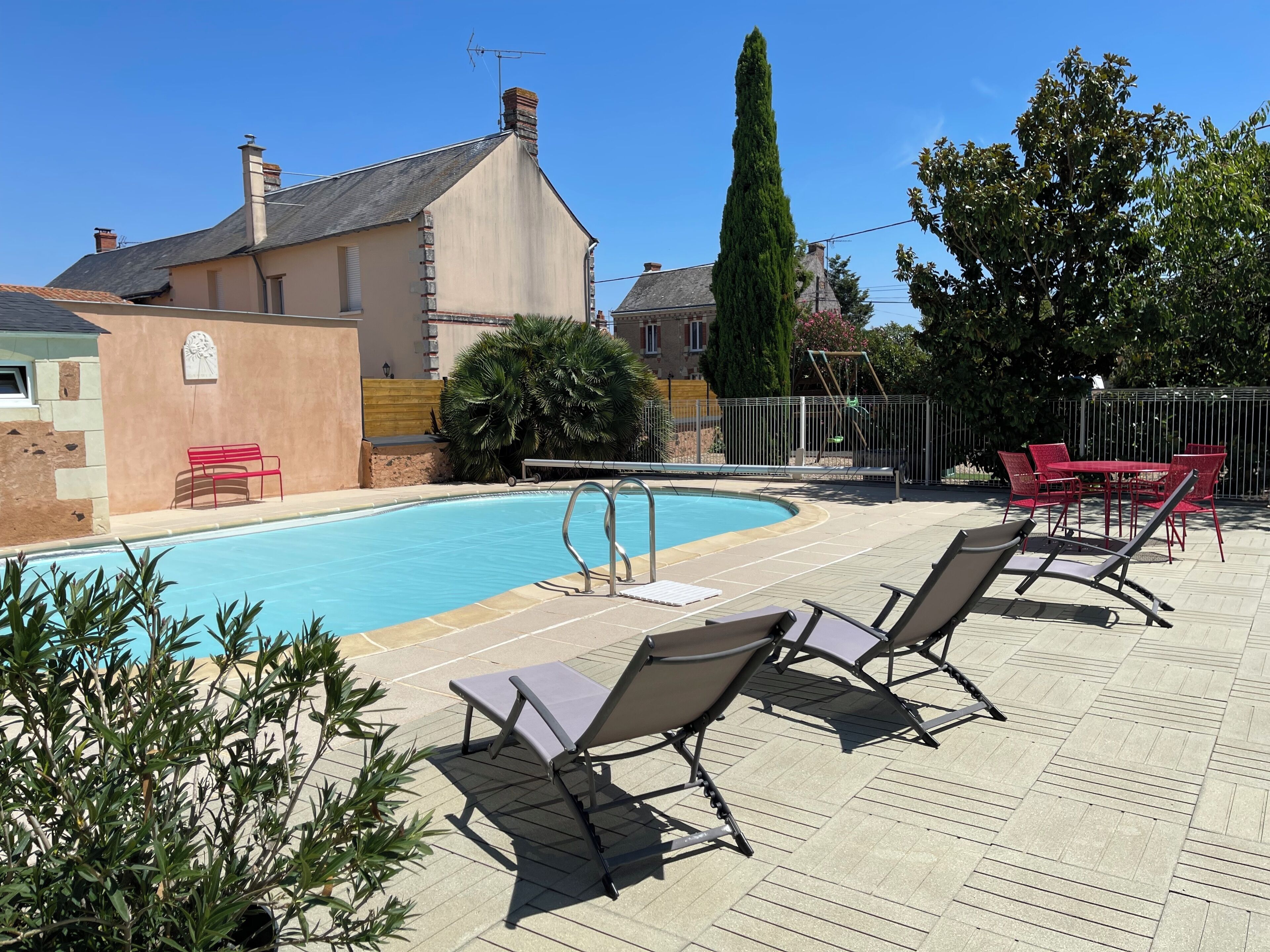 Une piscine entourée d’une clôture et de verdure, avec des chaises longues et une table à proximité.