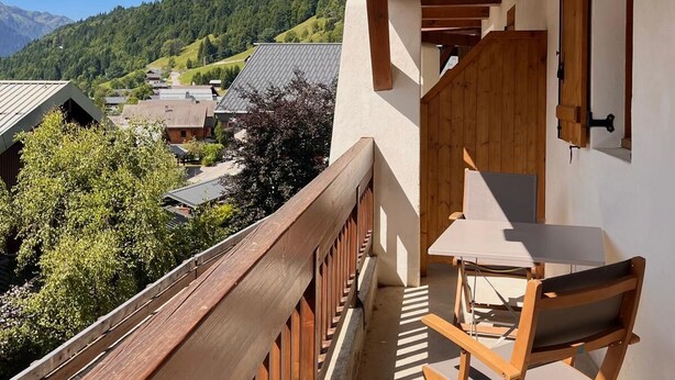 Ein Balkon mit Holzgeländer, einem Klappstuhl und Blick auf ein Dorf und Berge.