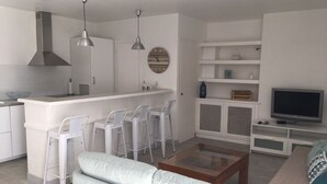 Ett modernt kök med vita sk åp och hängande lampor, en matplats med vita stolar, en soffa och en tv i vardagsrummet.