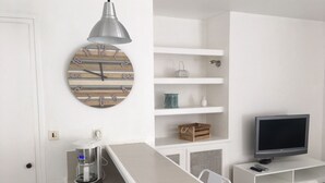 Ett modernt kök med en vit bänkskiva, en kaffemaskin, en klocka med siffror i trä, en hängande taklampa och en tv-apparat placerad på ett vitt skåp.