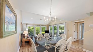 A spacious dining area with a glass-top table surrounded by white wicker chairs, a chandelier, and a view of the outdoors.
