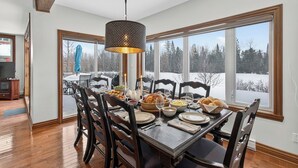 Comedor con una gran mesa de madera dispuesta para una comida, rodeada de sillas, y vista a un paisaje nevado a través de amplios ventanales.
