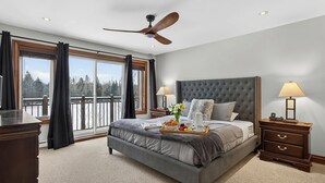 Un dormitorio con una cama grande, dos mesitas de noche con lámparas, un ventilador de techo y vistas al exterior a través de amplias ventanas.